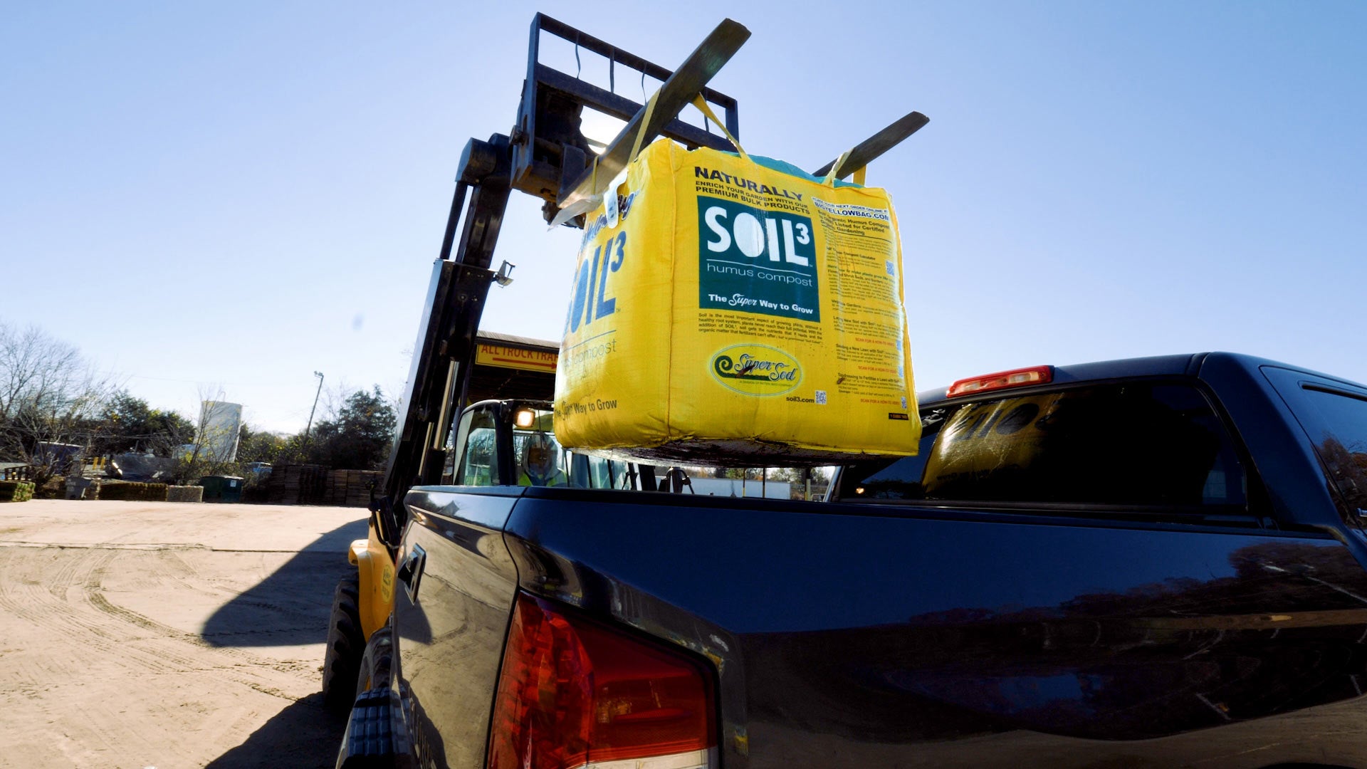 A pickup truck receiving a big yellow bag of soil cubed from a forklift